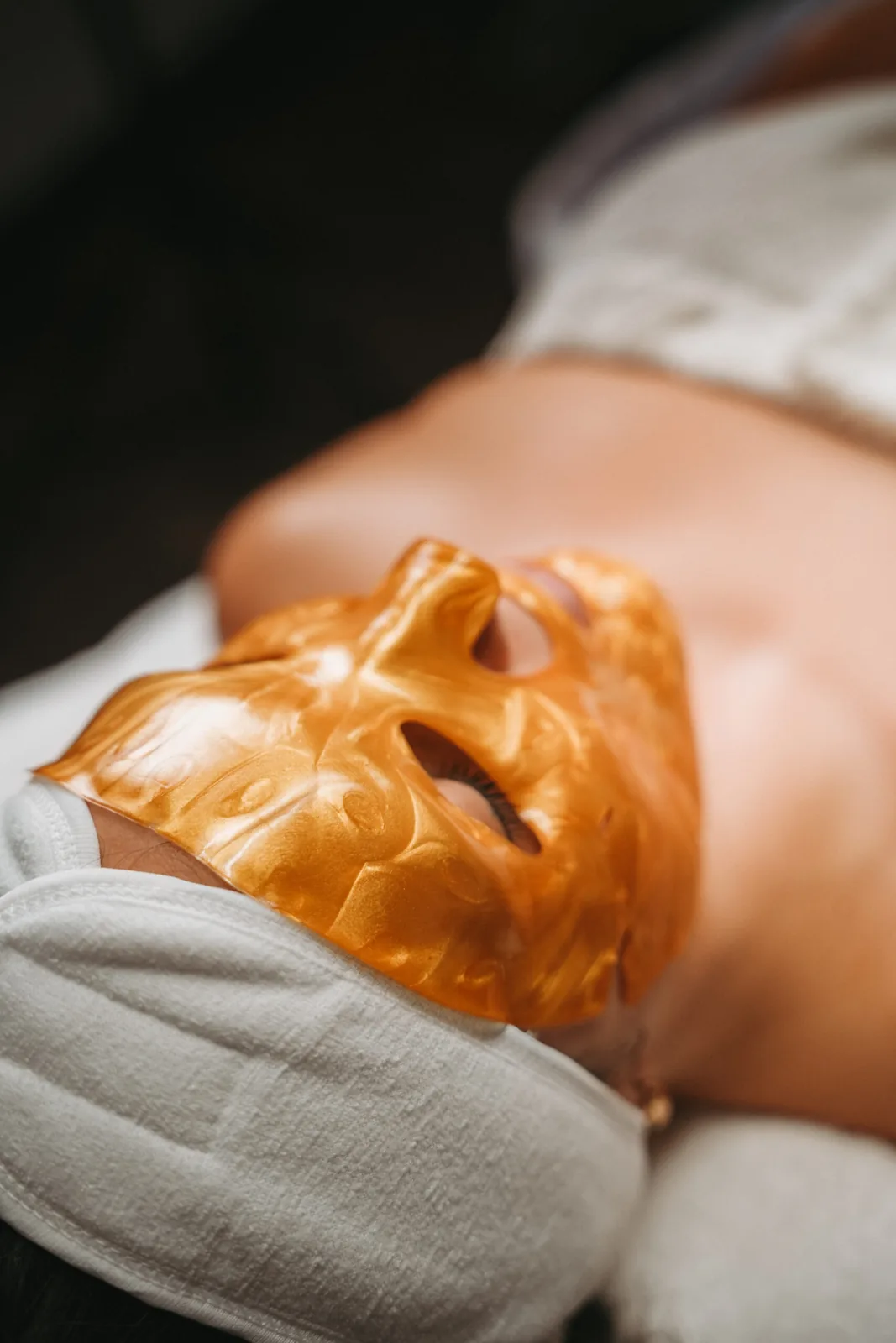 A woman reclining with a gold-colored facial mask and a white towel.