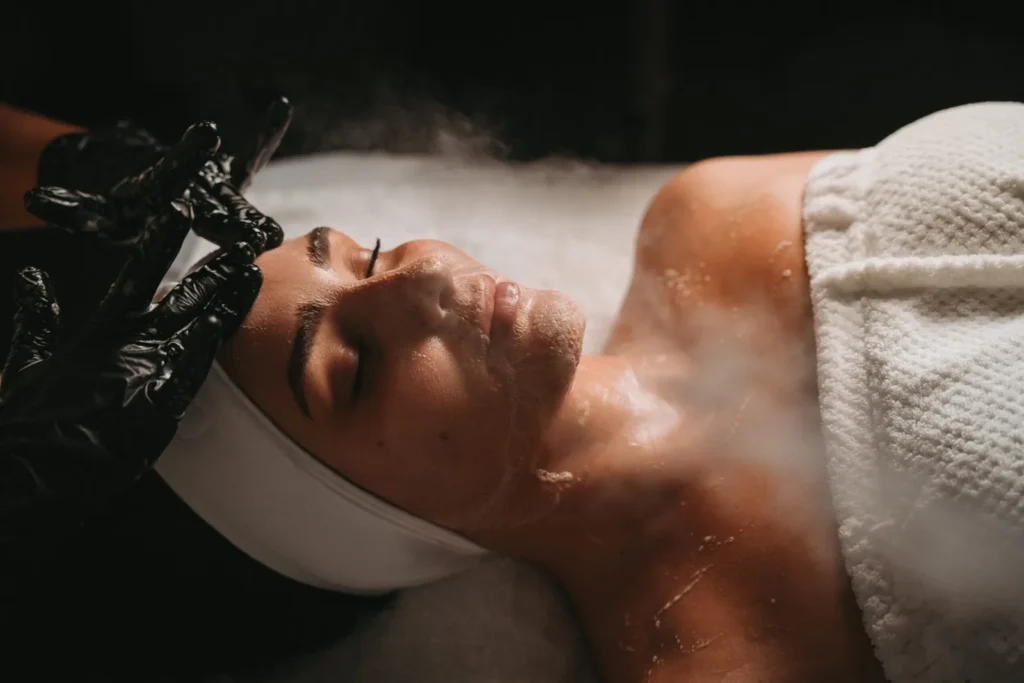 Gloved hands performing facial care while a woman rests on a treatment bed.